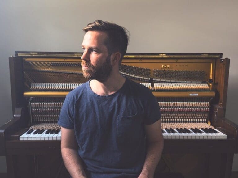 Bearded man in a blue t‑shirt sits in front of an open piano, looking to the left.