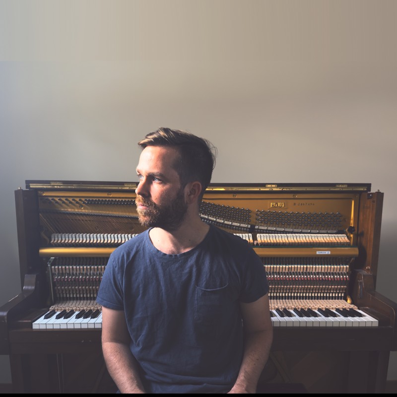 Bearded man in a blue t‑shirt sits in front of an open piano, looking to the left.