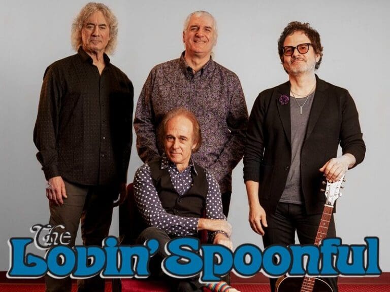 Four-member band photo: one seated front center with a patterned shirt, three standing around him, and a sunburst an acoustic guitar resting beside them. The Lovin' Spoonful logo is at the bottom left.