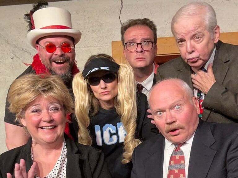 Six adults in quirky costumes posing for a group photo, some wearing wigs, hats, and sunglasses, smiling and making surprised faces.