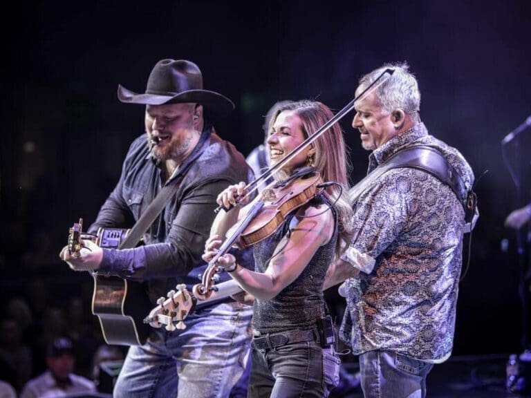 Three musicians on stage: a guitarist wearing a hat, a smiling violinist, and a fiddler performing to a crowd.