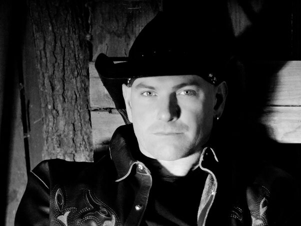 Man wearing a cowboy hat and embroidered leather jacket sits against a wooden wall, looking at the camera.