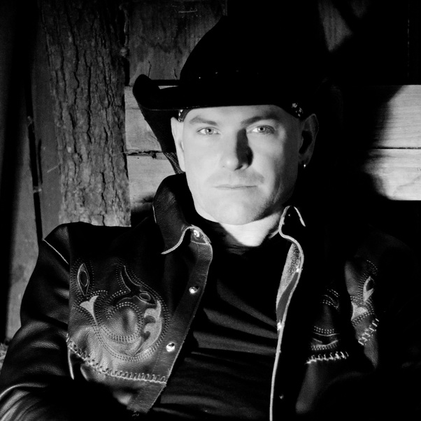 Man wearing a cowboy hat and embroidered leather jacket sits against a wooden wall, looking at the camera.