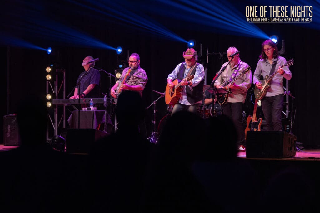 Live band on stage performing at night with blue stage lights and 'One Of These Nights' banner in the upper-right corner; tribute to The Eagles likely.