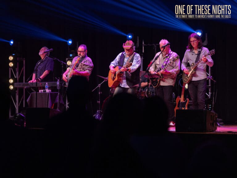 Live band on stage performing at night with blue stage lights and 'One Of These Nights' banner in the upper-right corner; tribute to The Eagles likely.