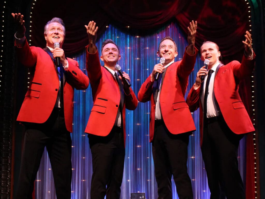 Four male singers in red blazers perform on a theater stage, each holding a microphone and raising a hand against a blue curtain backdrop.