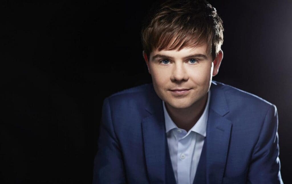 Portrait of a young man in a blue suit and white shirt, looking at the camera against a dark studio background.