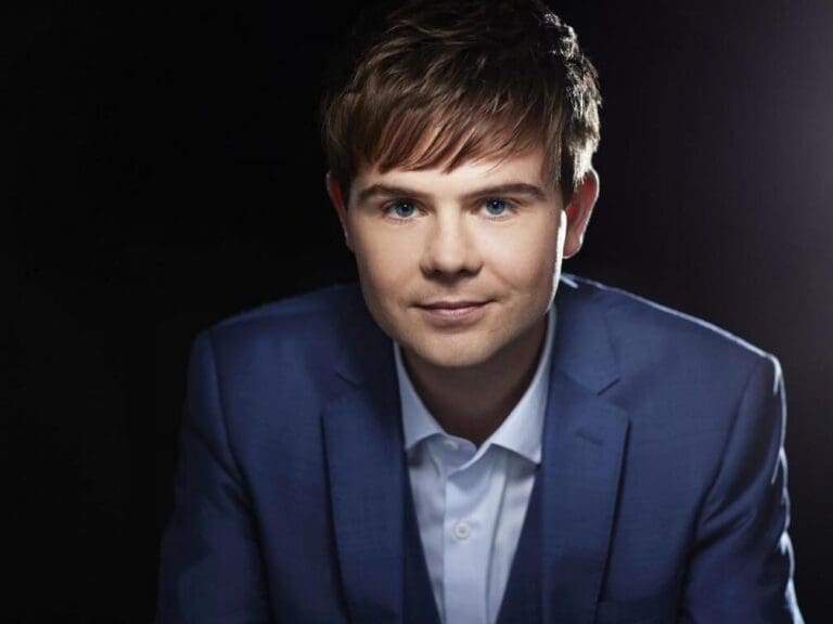 Portrait of a young man in a blue suit and white shirt, looking at the camera against a dark studio background.