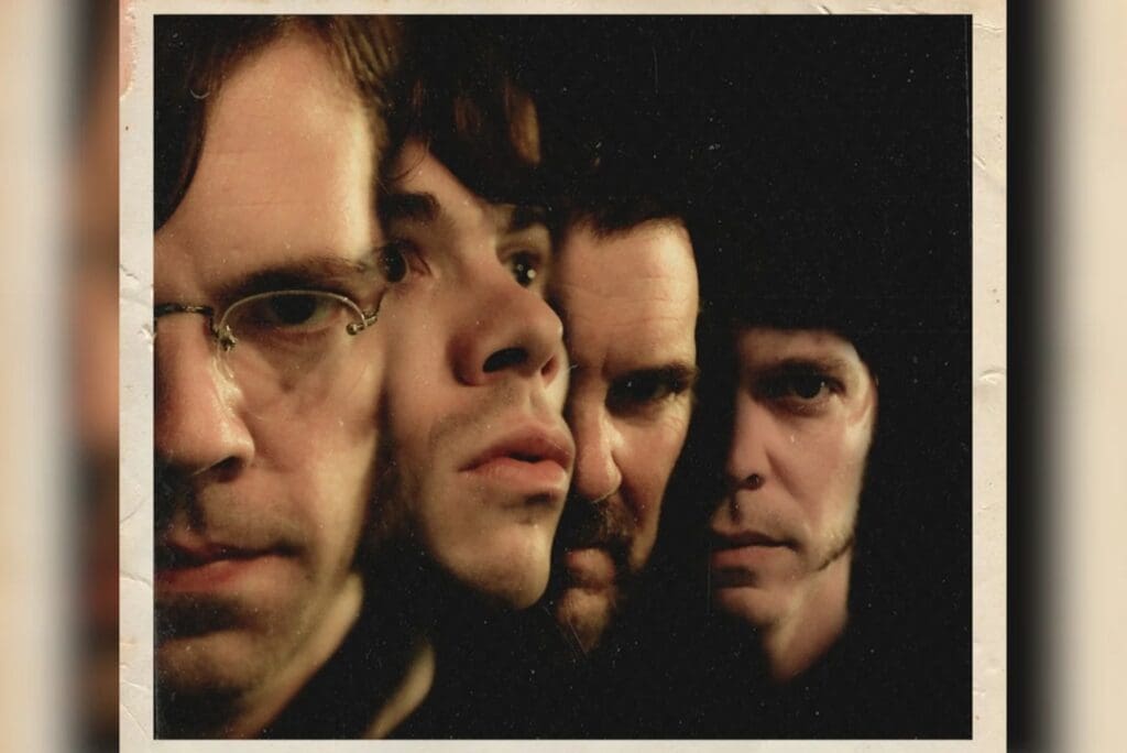 Close-up group portrait of four men with serious expressions, tightly framed in a dark, moody setting.