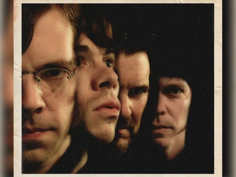 Close-up group portrait of four men with serious expressions, tightly framed in a dark, moody setting.